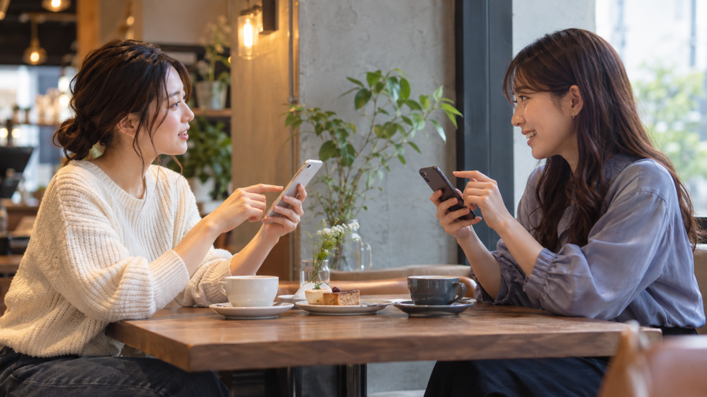 カフェでそれぞれのスマートフォンを見ながら予定を相談する友人